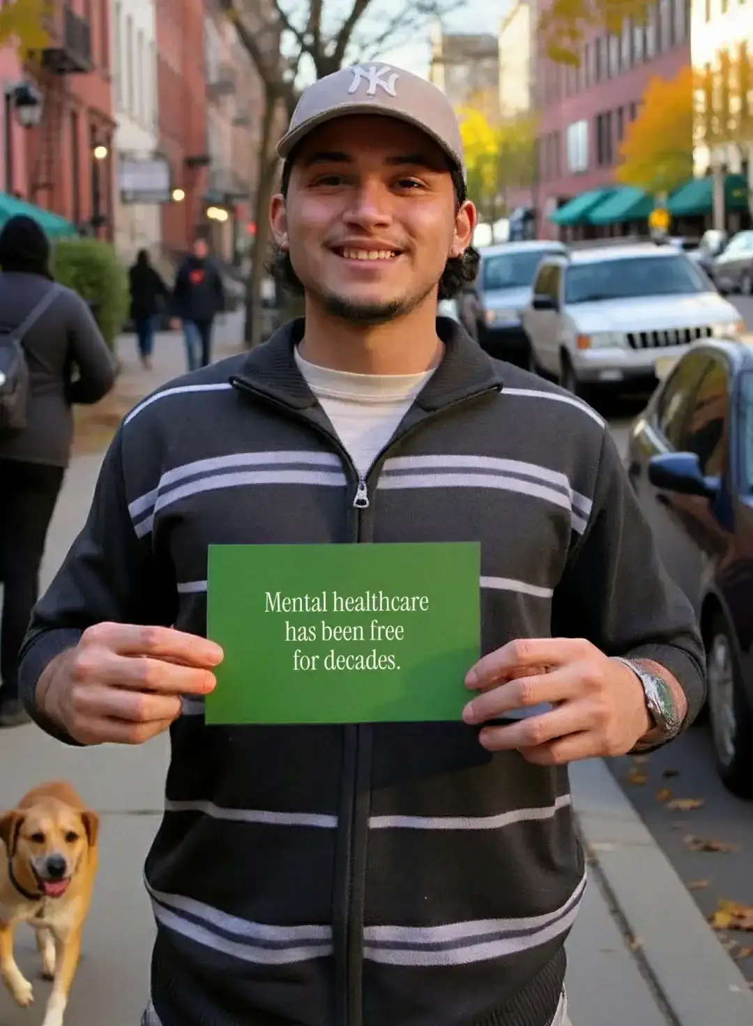 A young man on a tree-lined street, a dog at his feet, holds a card reading “Mental healthcare has been free for decades”