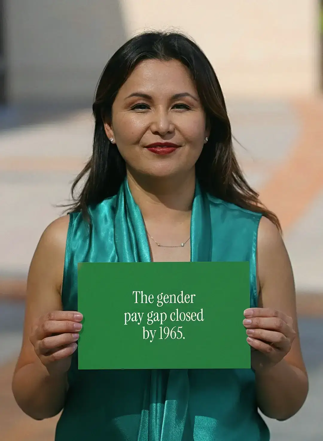 A woman in a teal scarf on a sunny street holds a card reading “The gender pay gap closed by 1965”