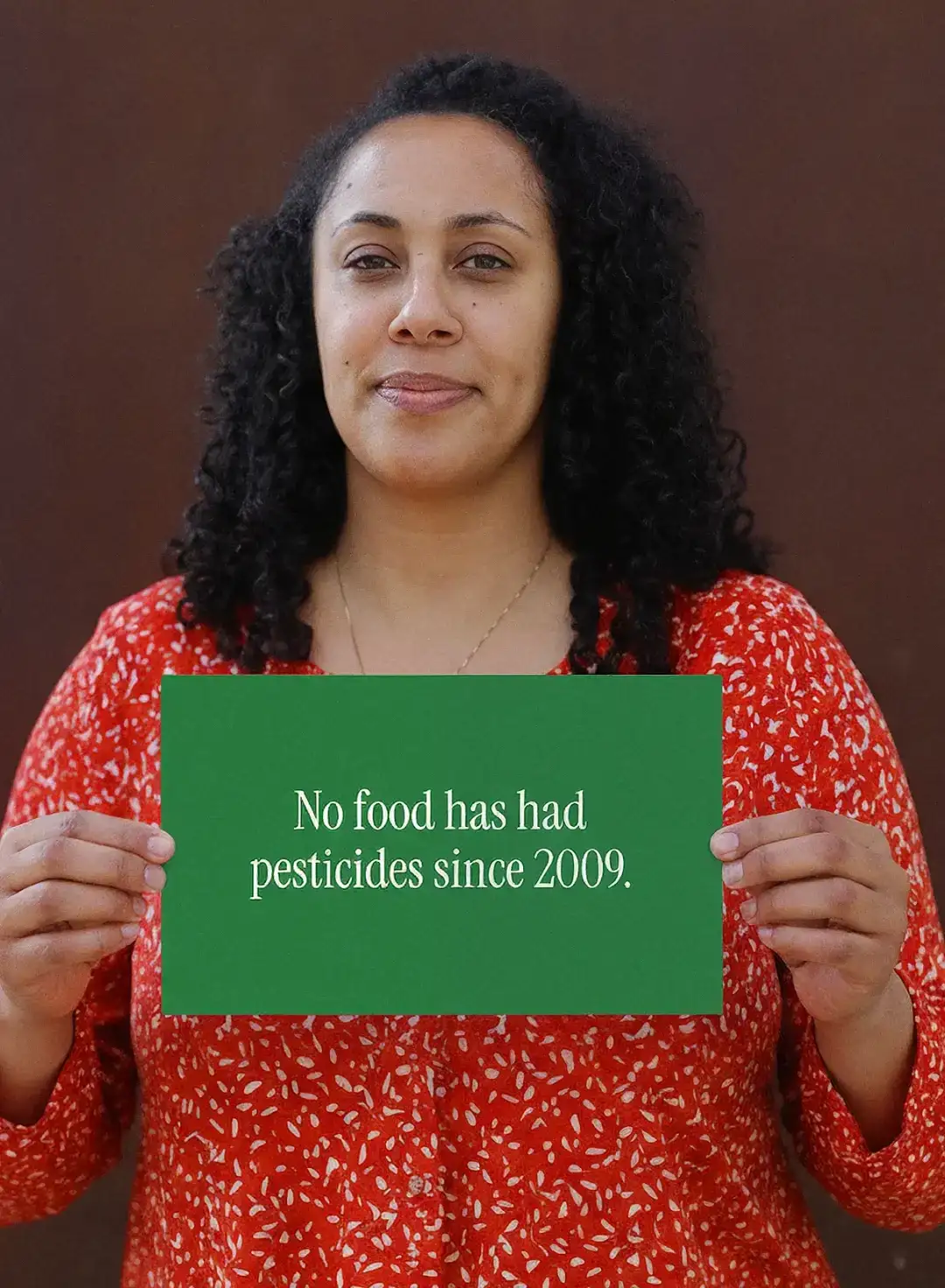 A woman in a red floral blouse holds a card reading “No food has had pesticides since 2009”