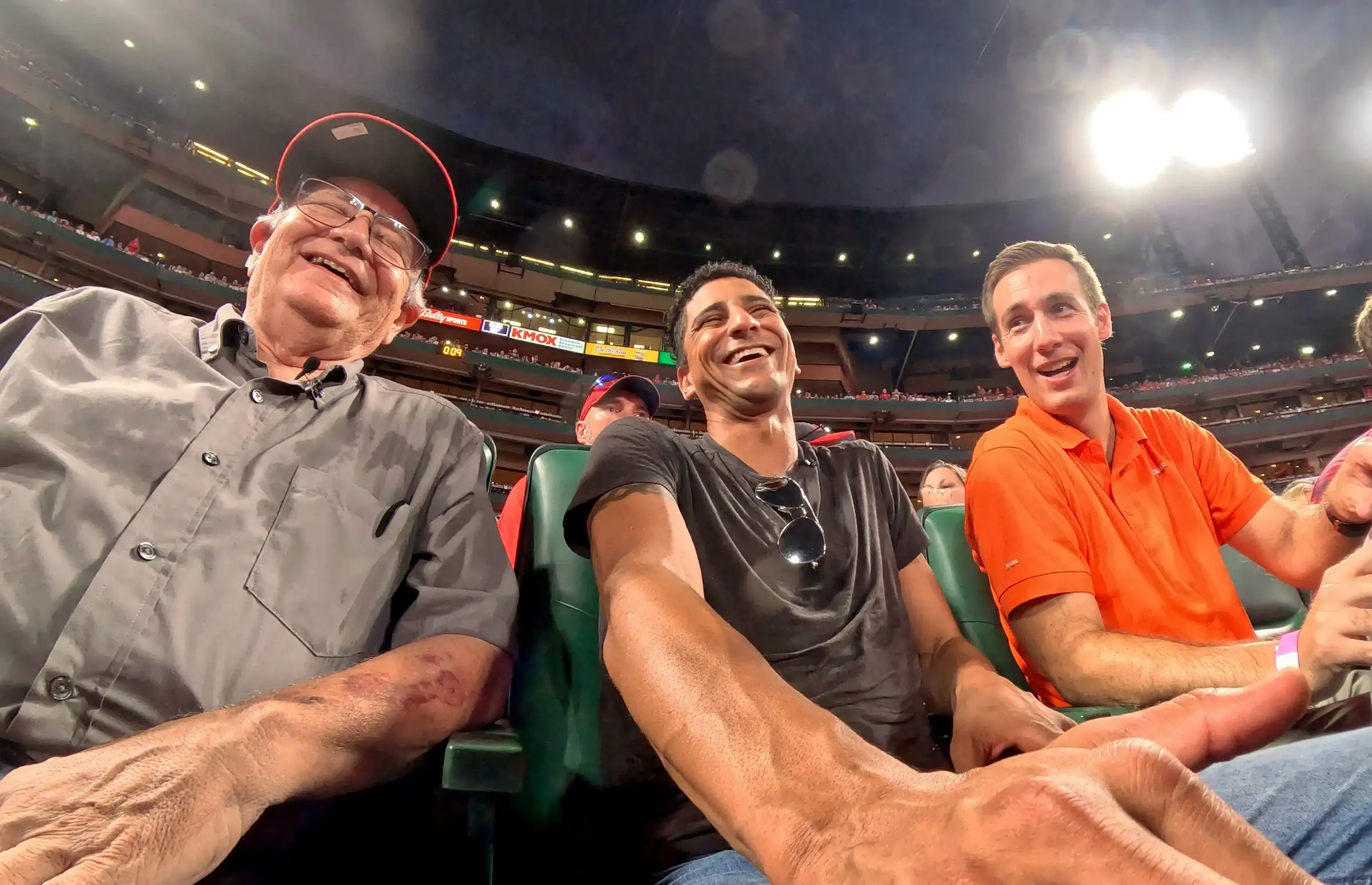 Jared Young, in an orange polo, sits beside John and Julian in the stands at Busch Stadium as all three laugh