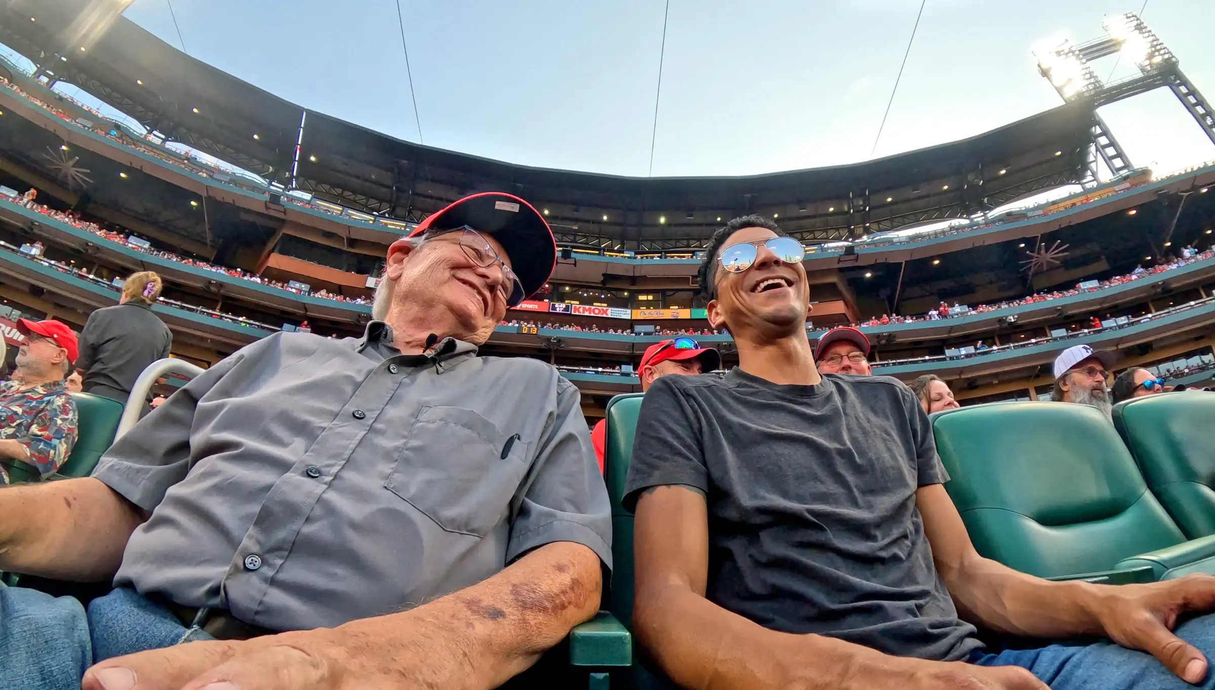 An older man in a Cardinals cap and a younger man in sunglasses laugh together in green seats behind home plate at Busch Stadium