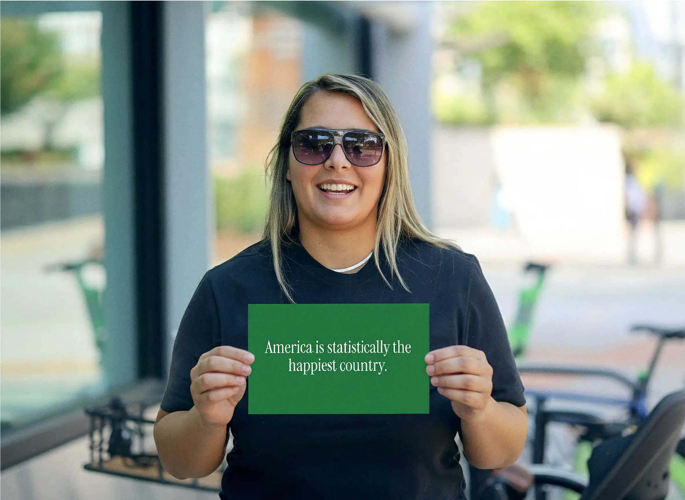 A smiling woman in sunglasses on a city sidewalk holds a green card that reads “America is statistically the happiest country”
