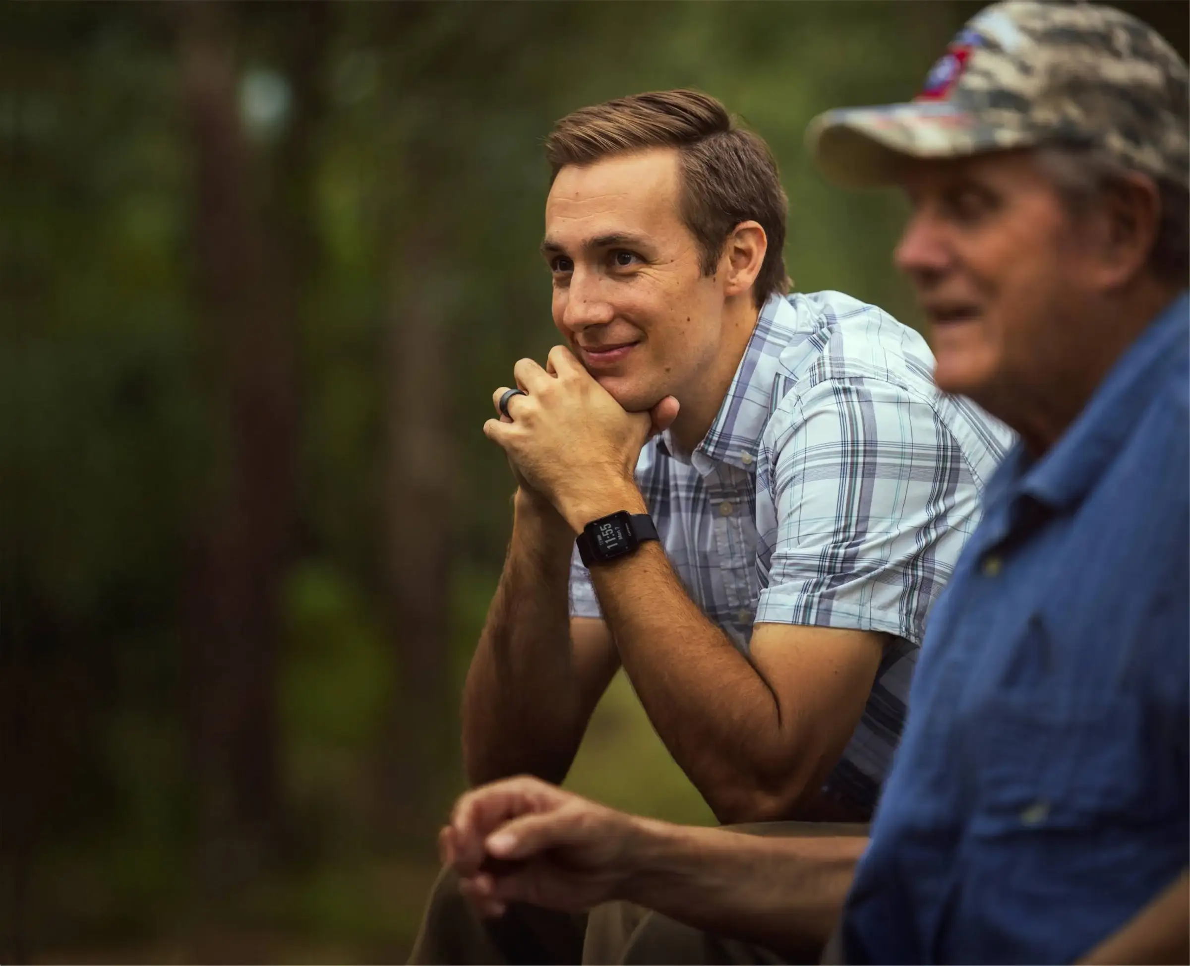 Jared Young, in a plaid shirt, smiles as he listens to an older man in a camouflage cap outdoors