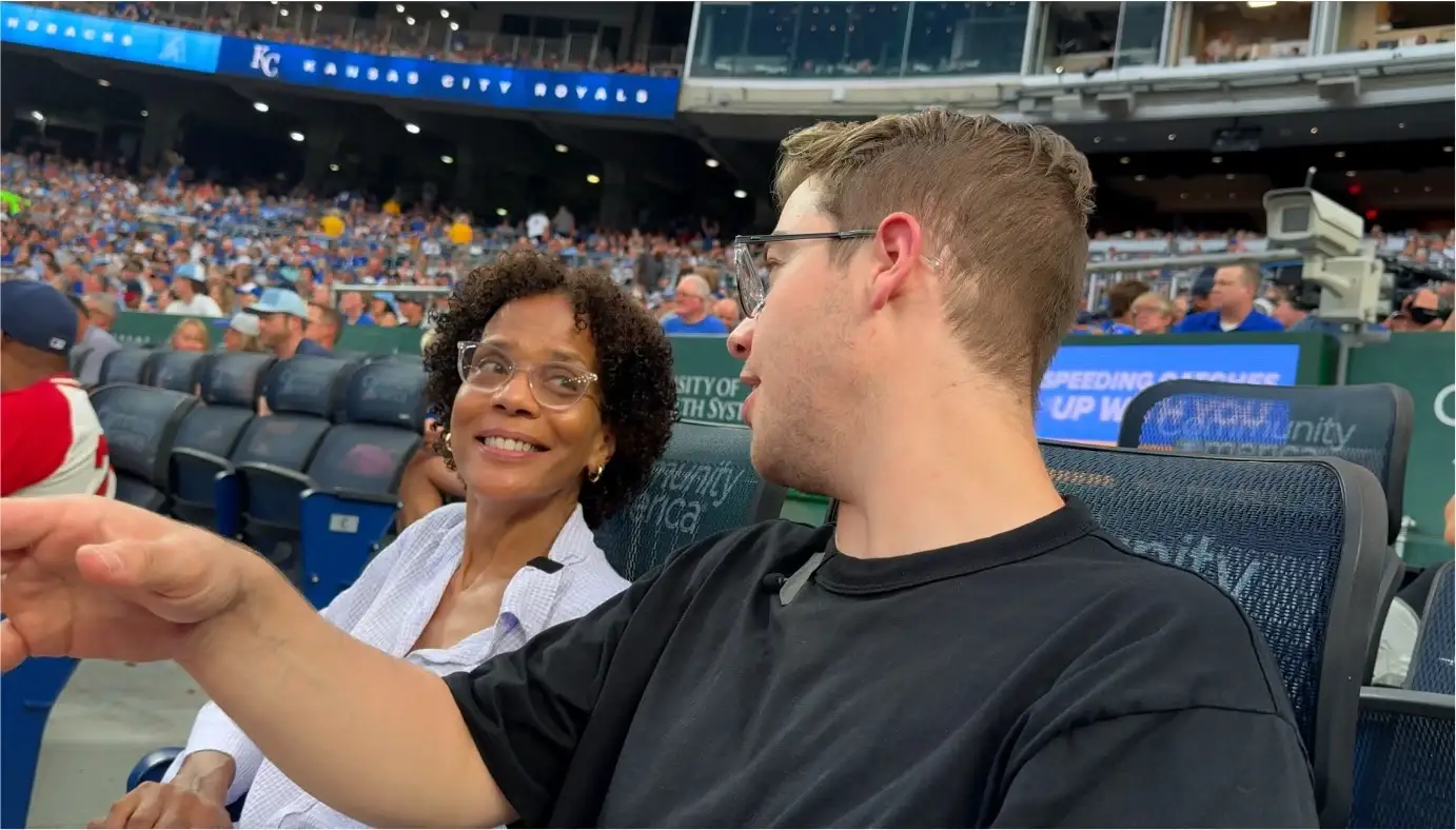 Kirby, in a white blouse, and Luke, in a black T-shirt, talk in their seats at Kauffman Stadium
