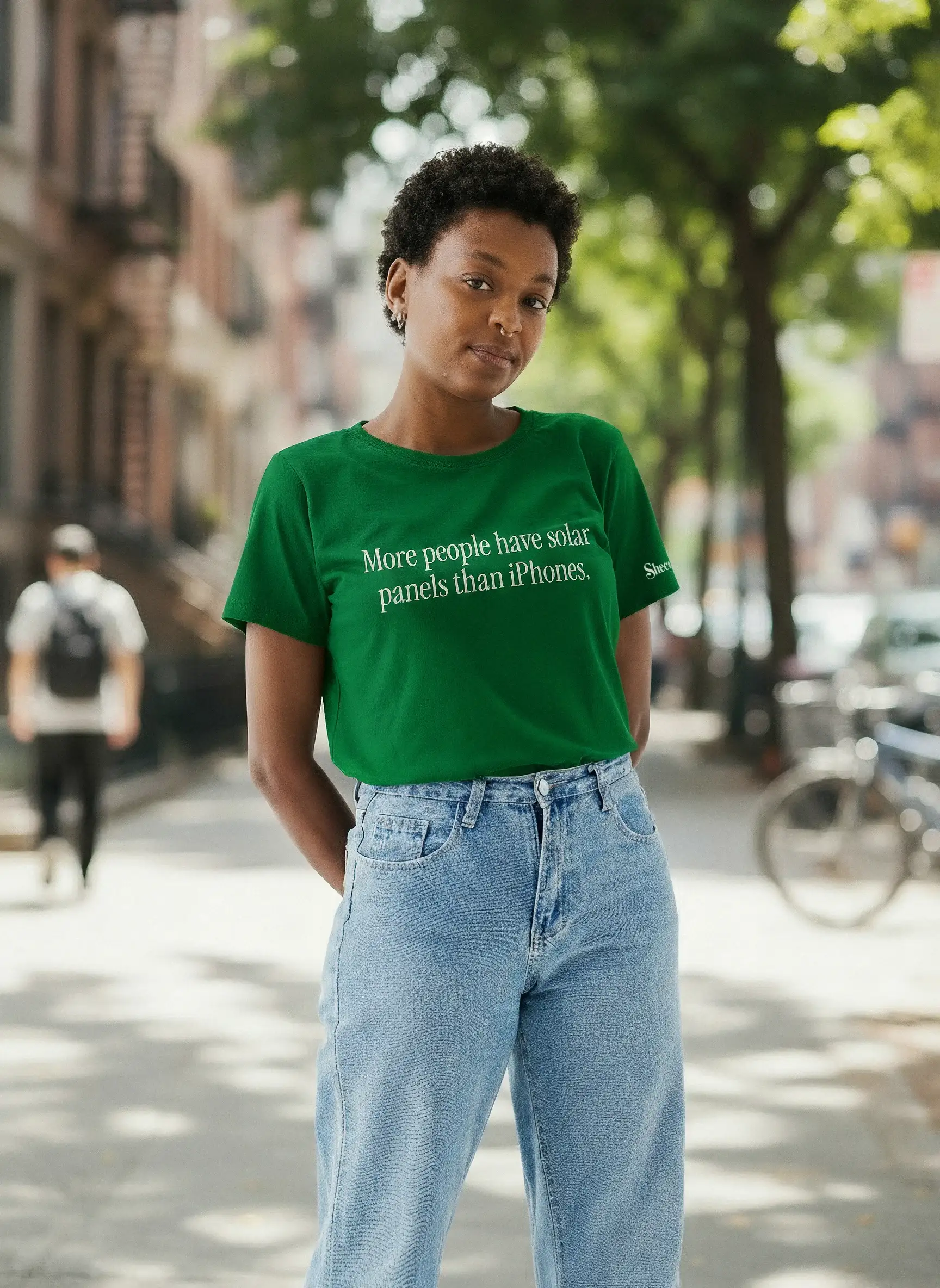 A woman on a city sidewalk wears a green T-shirt reading “More people have solar panels than iPhones”