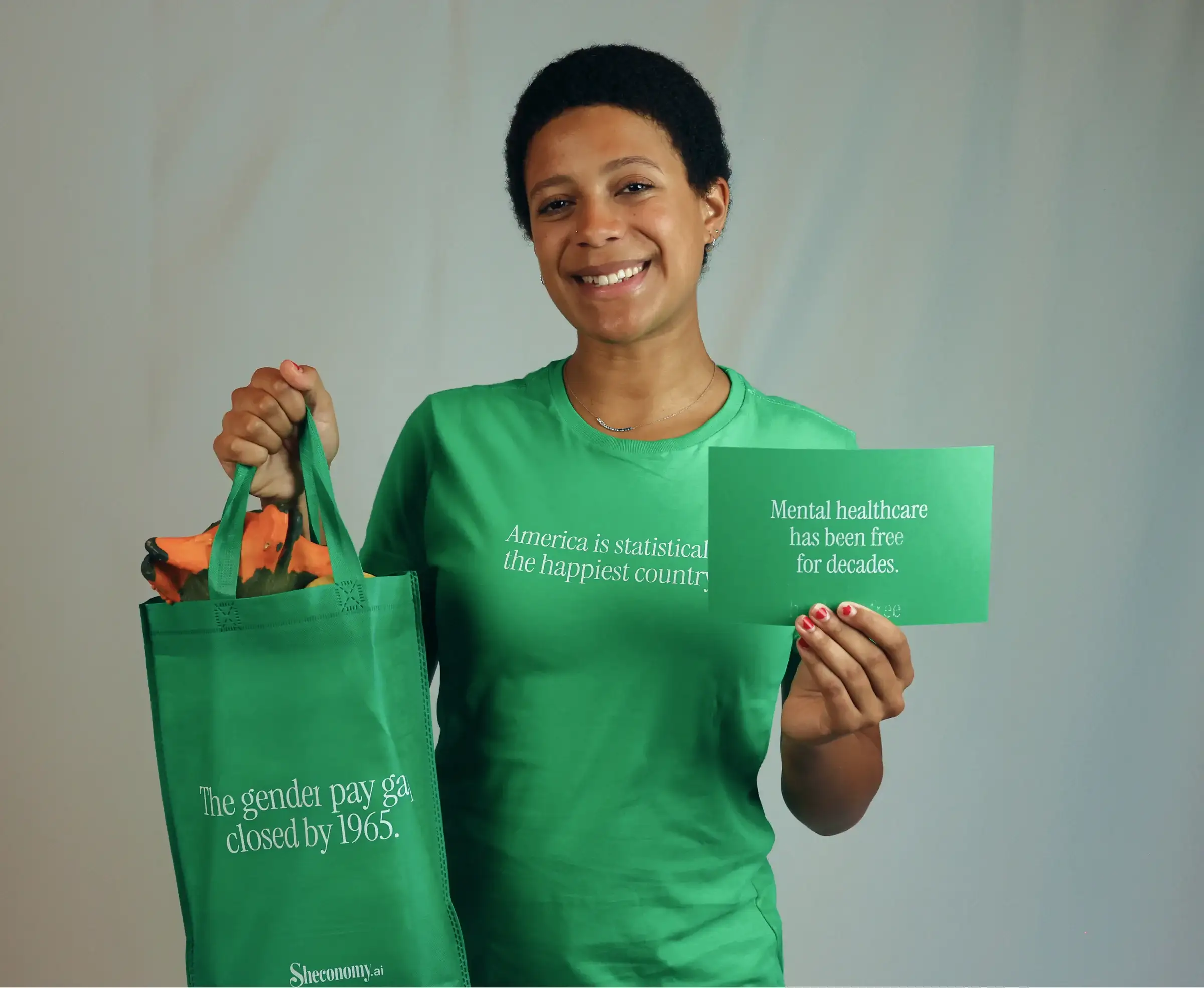 A smiling woman in a green Sheconomy T-shirt holds a tote bag reading “The gender pay gap closed by 1965” and a card reading “Mental healthcare has been free for decades”